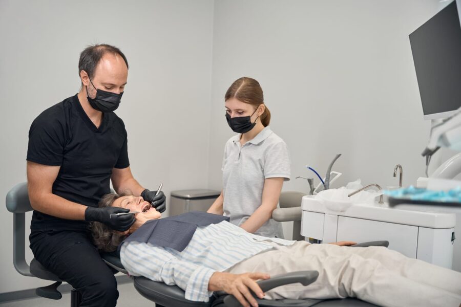 Middle-aged doctor treats an elderly ladys teeth, a young assistant helps him with his work